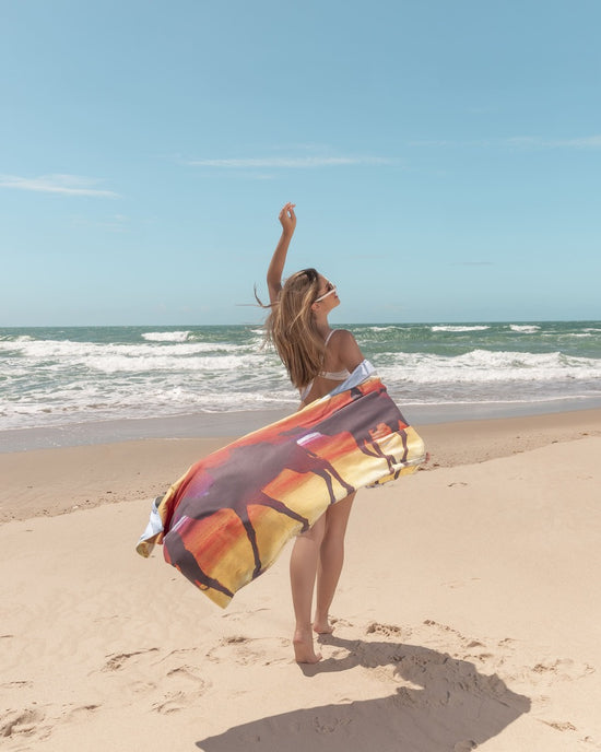 Model with Broome Microfibre Artistic Towel at the beach. The towel shows Broome’s famous camel ride at the beach during sunset and the full moon in the sky being reflected by the wet coastal sand. Art inspired by the red desert and clear blue waters.