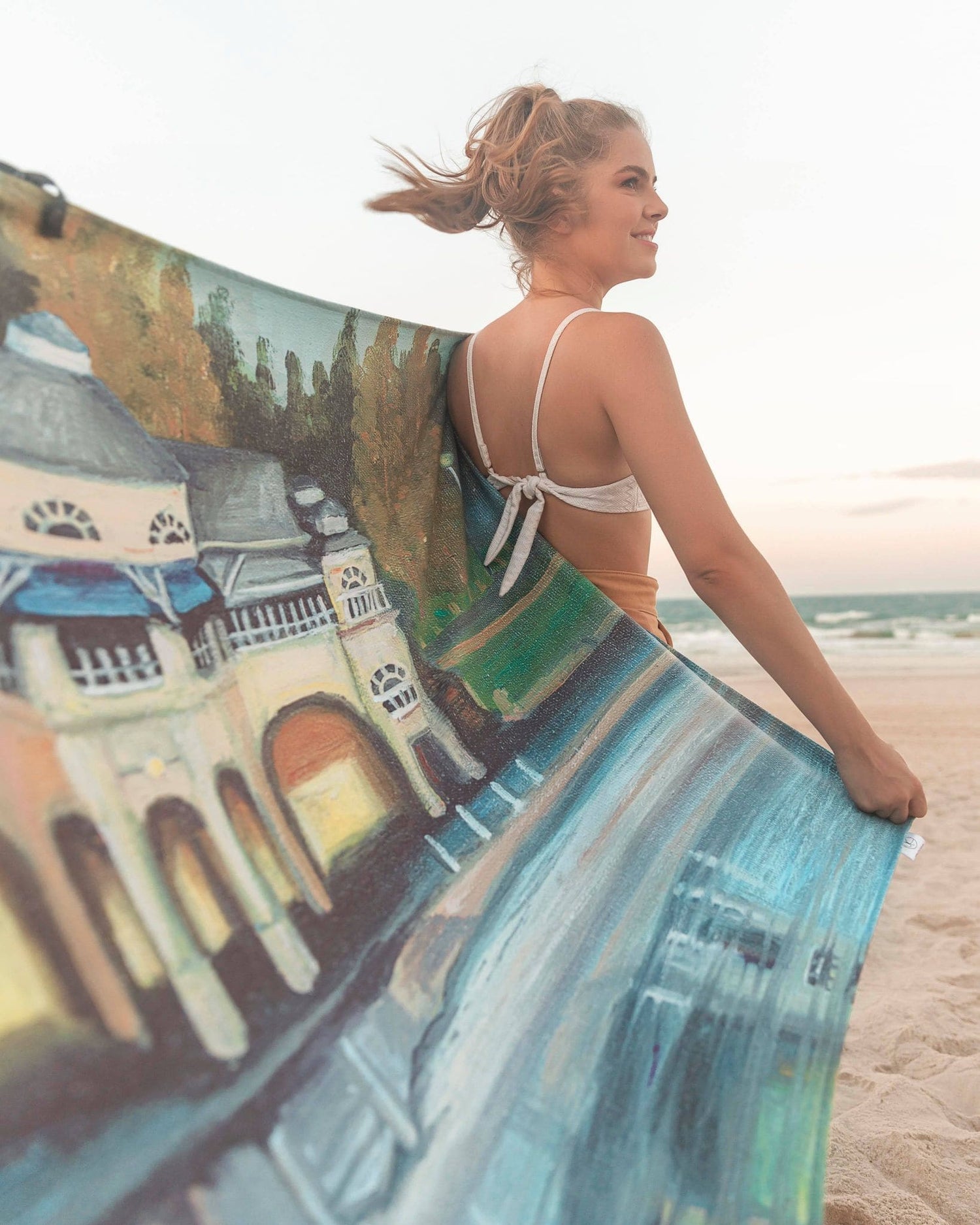 Female model holding Cottesloe Beach microfibre artistic towel. The towel shows the ocean, the Pavilion and trees behind it. Large size, 170cm x 95cm, soft touch, compact and sand free beach towel. An Aussie-inspired art showcasing magnificent landmarks and the Aussie lifestyle