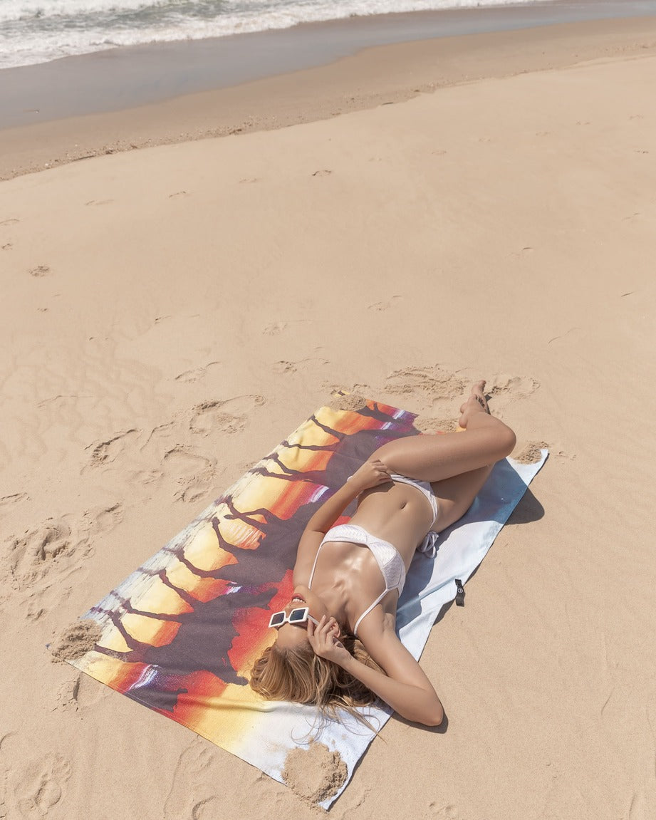 Model tanning at the beach lying on Broome microfibre artistic towel. The towel shows Broome’s famous camel ride at the beach during sunset and the full moon in the sky being reflected by the wet coastal sand. An art also inspired by the contrast of the red desert with the clear blue waters of the ocean. Large size, 170cm x 95cm, soft touch, compact and sand free beach towel. An Aussie-inspired art showcasing magnificent landmarks and the Aussie lifestyle.