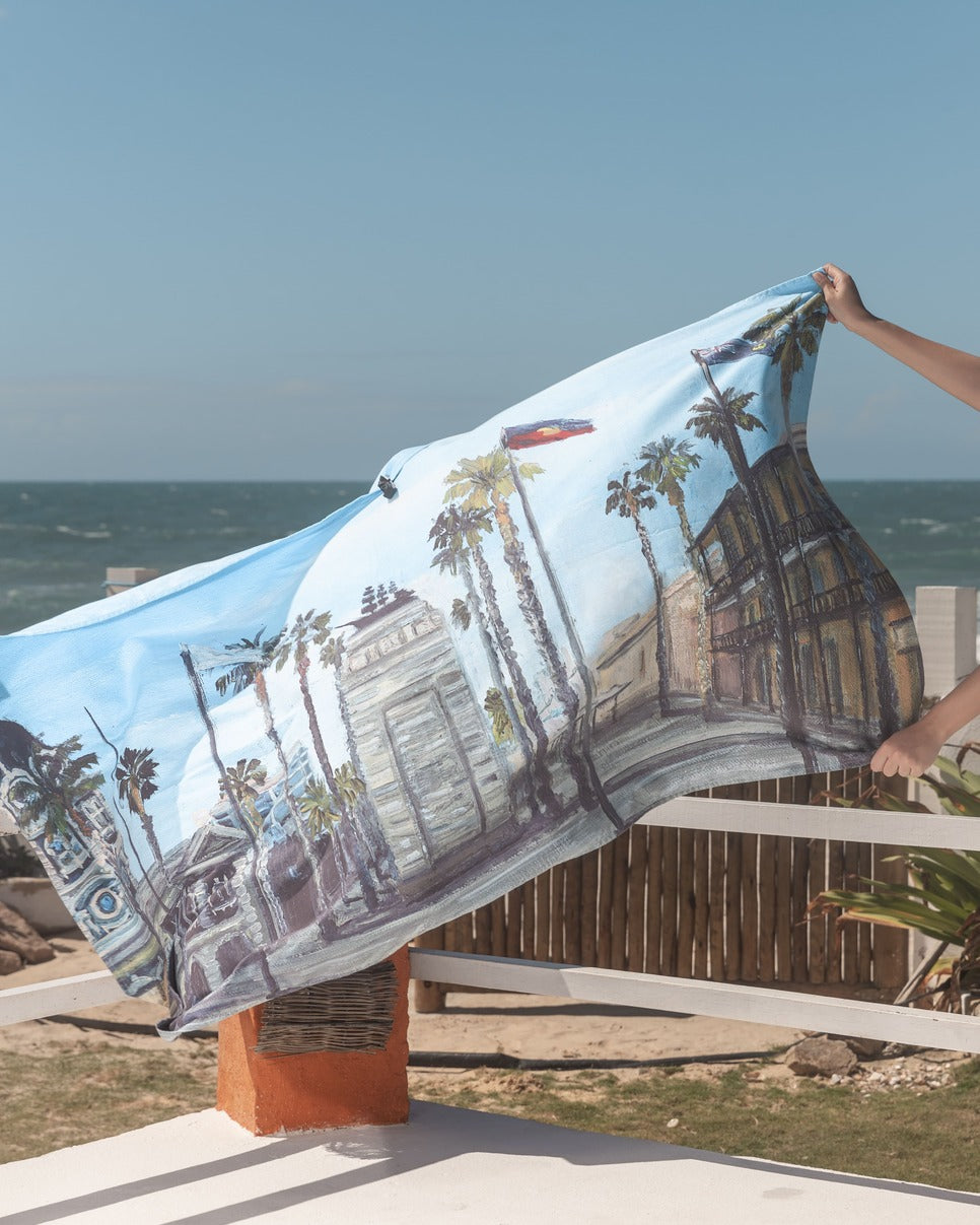 Glenelg Microfibre Artistic Towel being hold and the ocean on the background. Our artistic towel shows the iconic Moseley square located in Glenelg, Adelaide. The artwork emphasis the Pioneer Memorial, the abundant palm trees, and the historical buildings. Large size, 170cm x 95cm, soft touch, highly absorbent and quick-drying, compact towel. An Aussie-inspired art showcasing magnificent landmarks and the Aussie lifestyle.